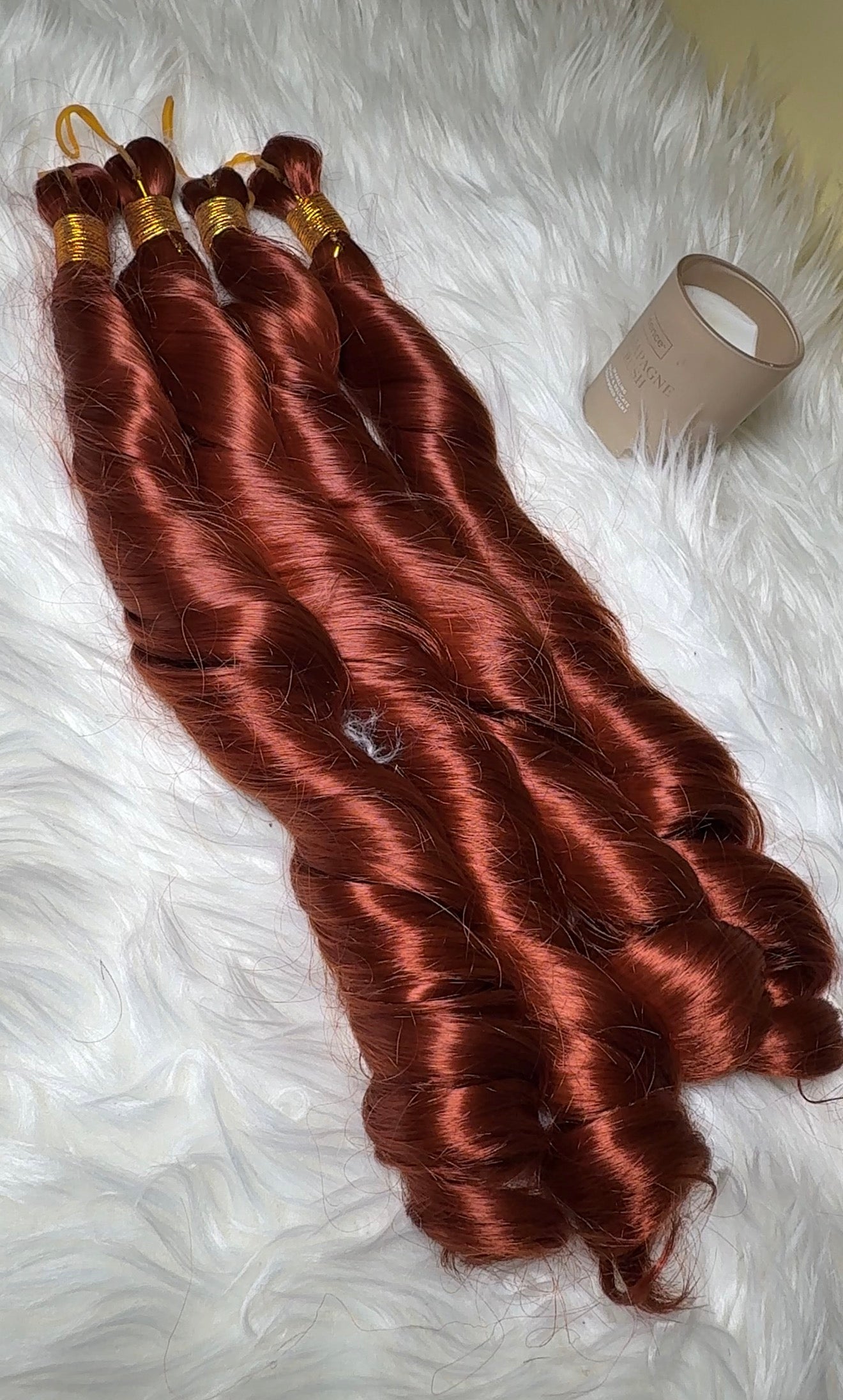 Four bundles of wavy coppery auburn hair extensions laid out on a white faux fur surface, with a beige plastic container visible in the upper right.