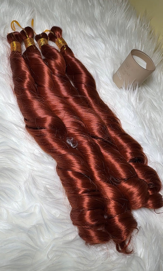 Four bundles of wavy coppery auburn hair extensions laid out on a white faux fur surface, with a beige plastic container visible in the upper right.